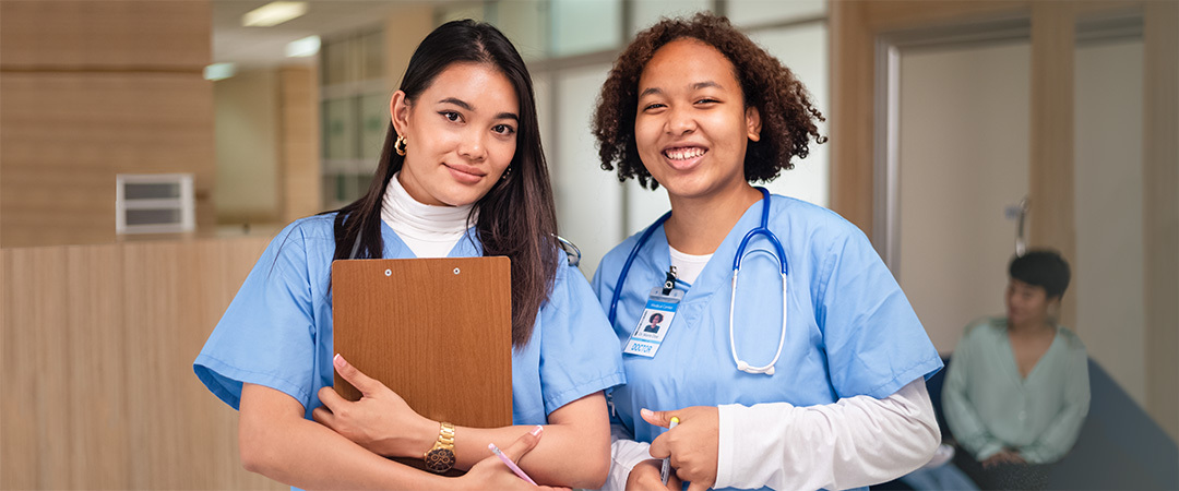Two young nurses holding clipboards talking in a hallway